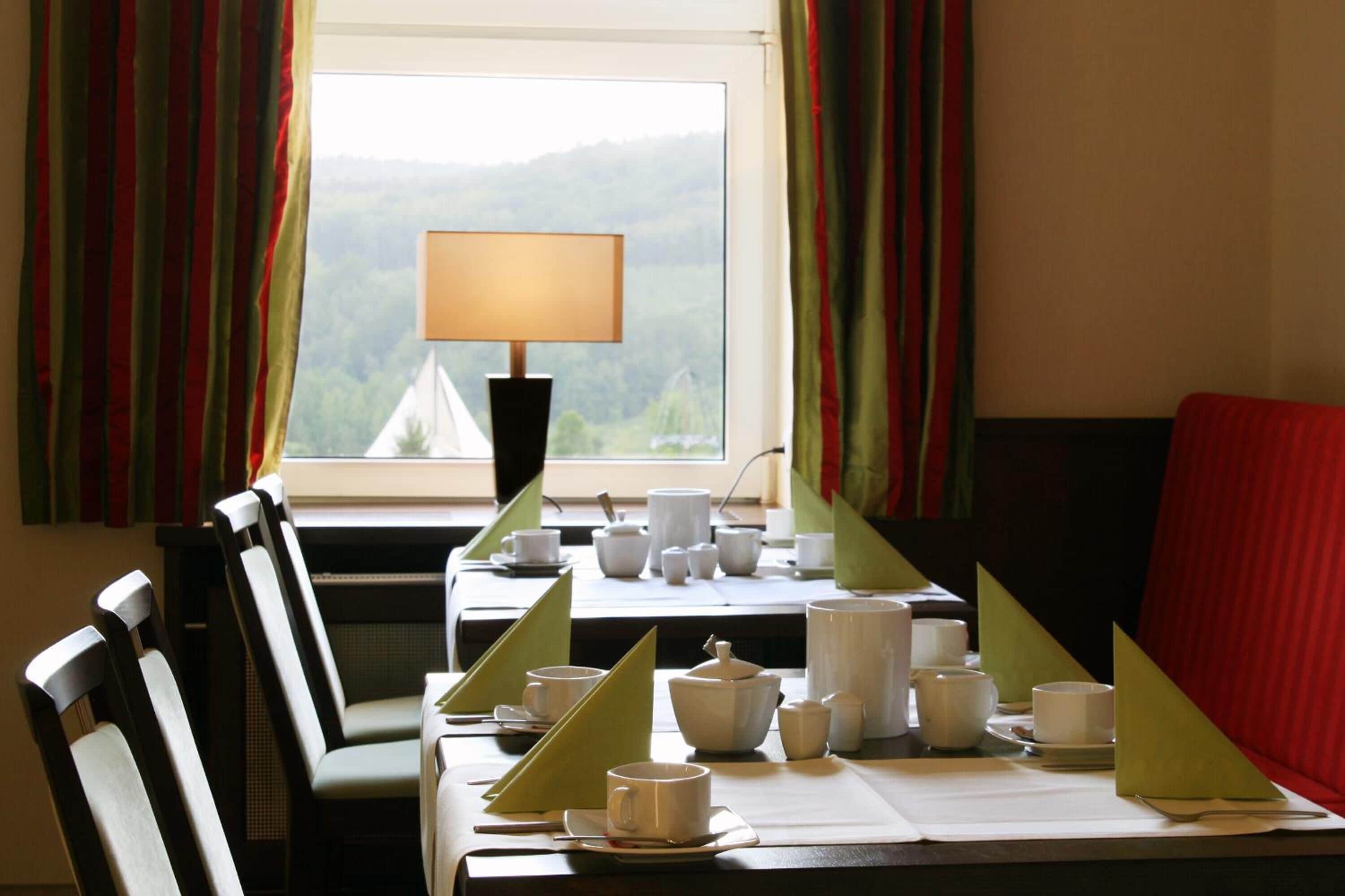 Un comedor con una mesa dispuesta para dos, una lámpara y vistas a un bosque por la ventana.