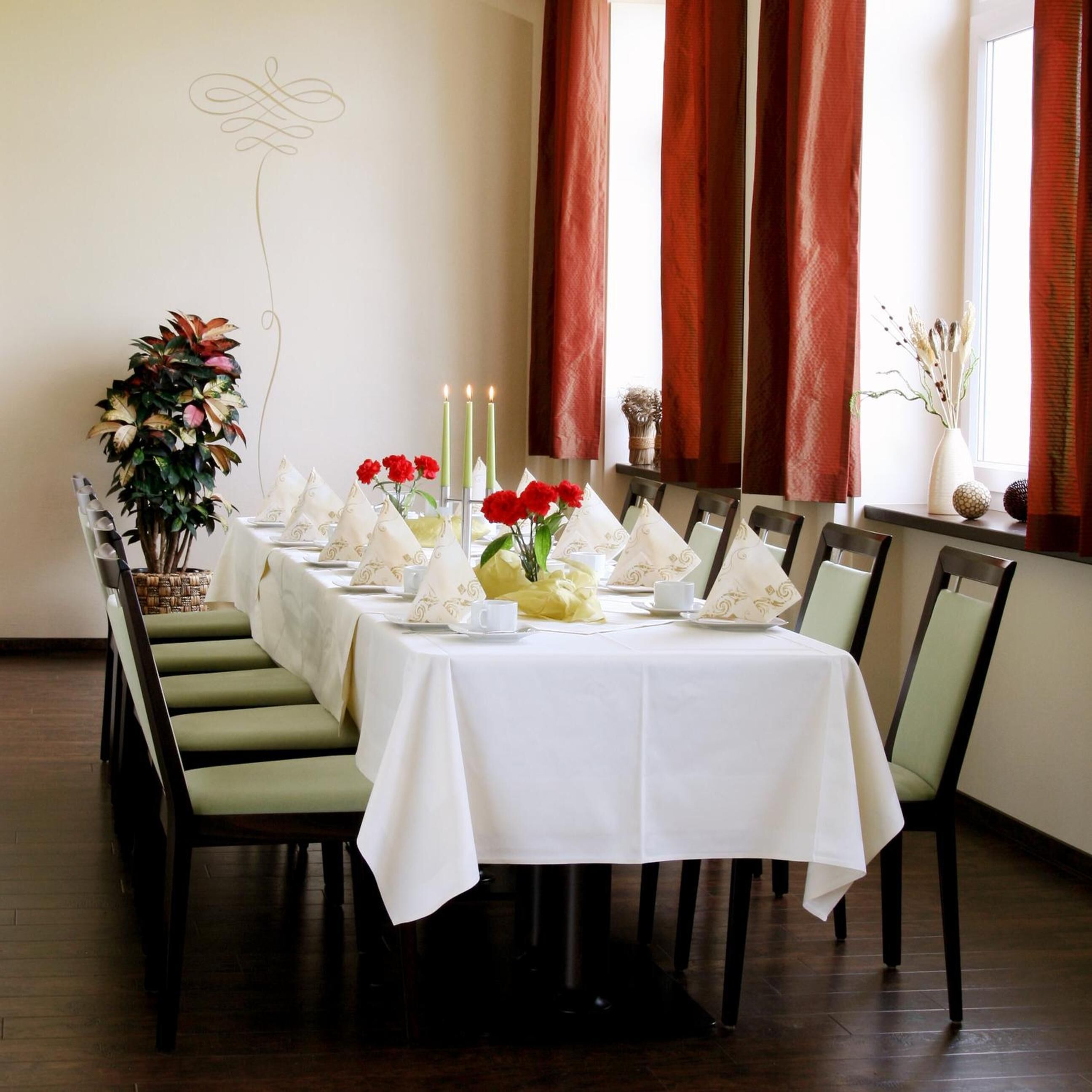 A dining area with a white table set for six, green upholstered chairs, and a potted plant on the left.