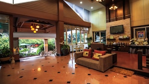 A spacious lobby with a high ceiling, a seating area with a sofa and chairs, a TV mounted on a stone wall, and a large window providing a view of the outdoors. - Kwanruen Resort (Nakhon Ratchasima)