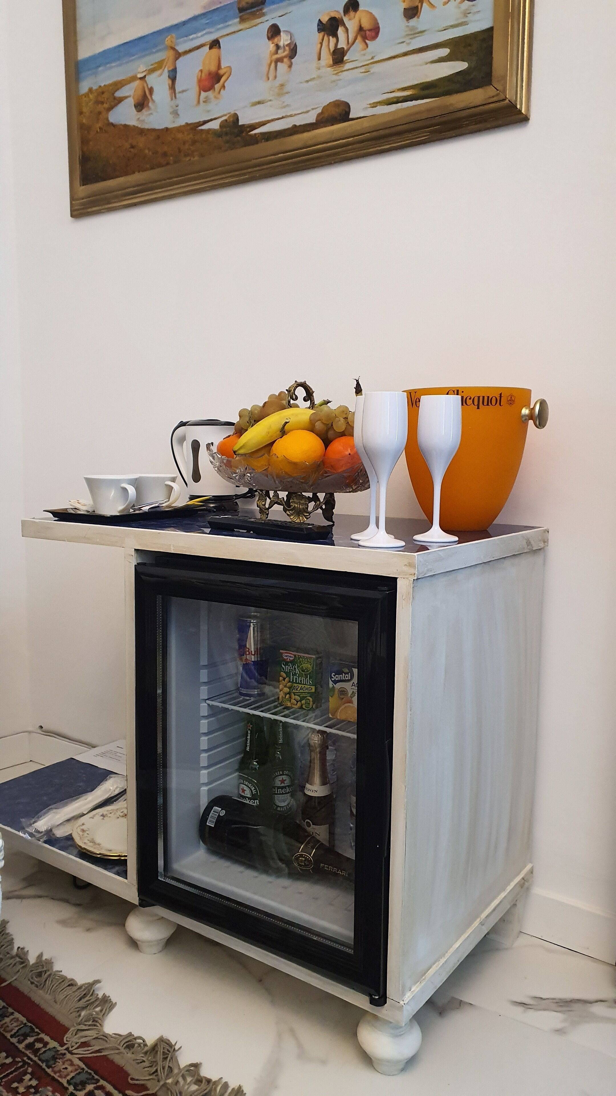 A small bar cart with a refrigerator, a bowl of fruit, and two wine glasses.