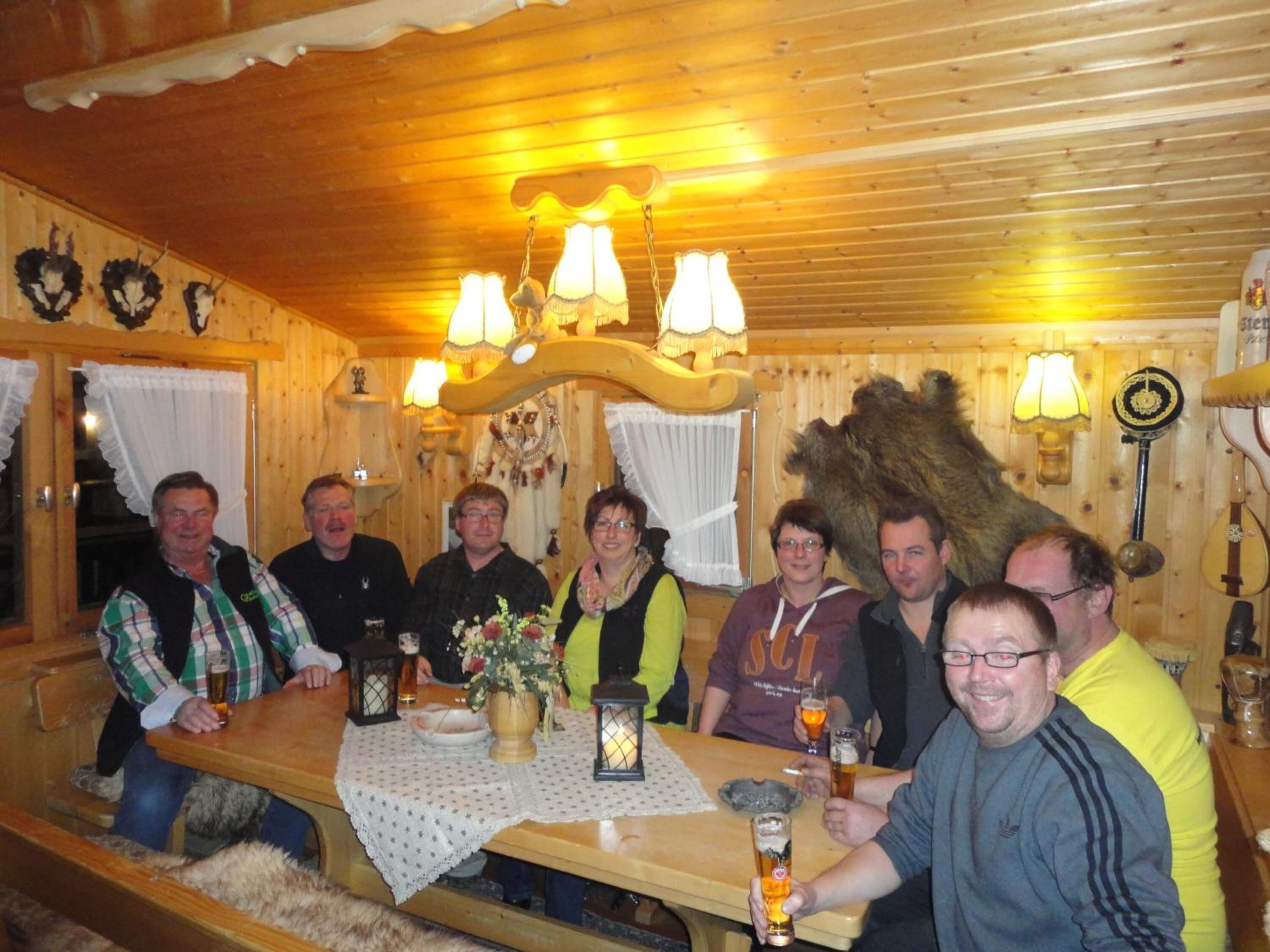 Eine Gruppe von Personen sitzt an einem Holztisch in einem gemütlich beleuchteten Raum mit Holzwänden und -decke, der einen rustikalen Charme versprüht, darunter ein großer Fellteppich und Wandlampen.
