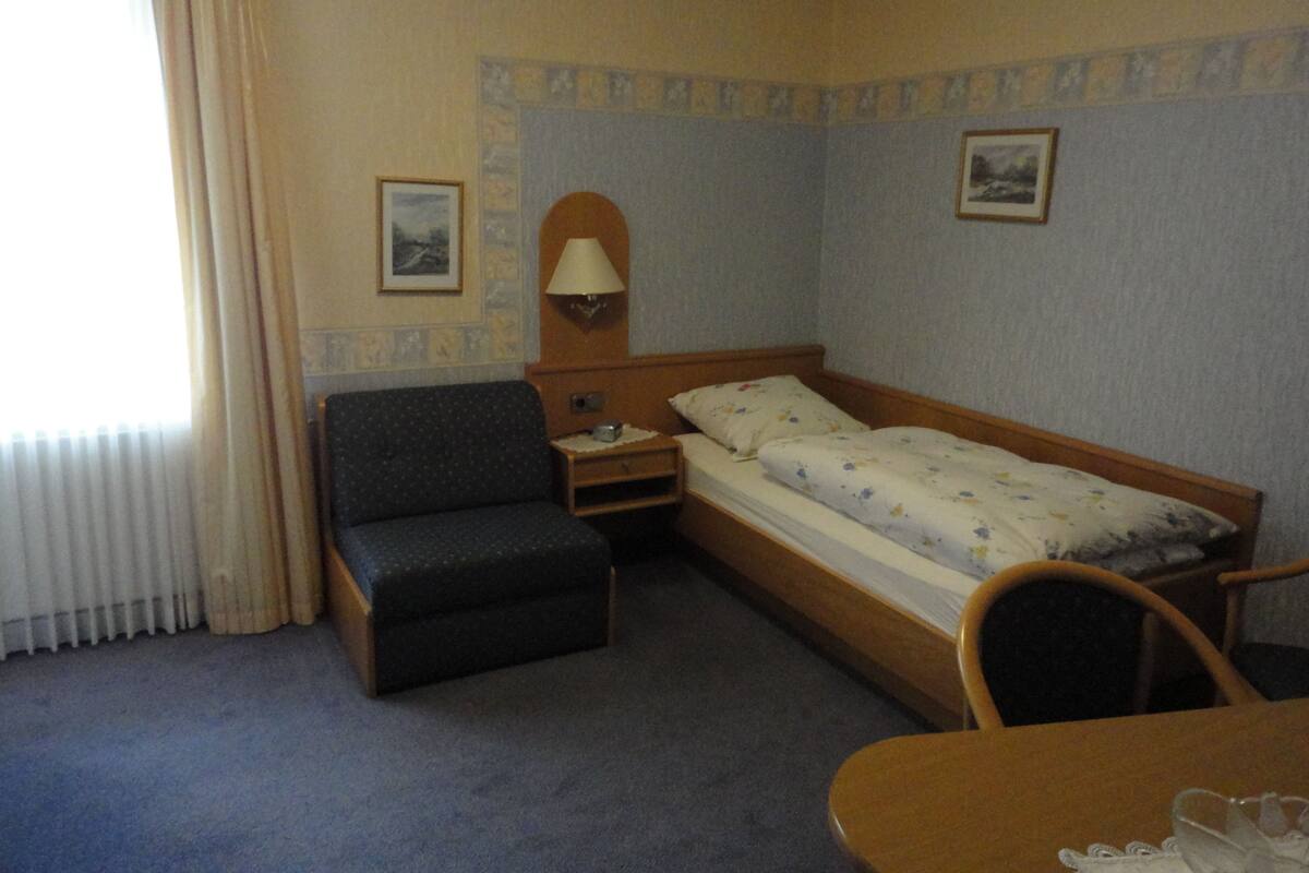 Ein Hotelzimmer mit einem Einzelbett, einem Sessel, einem kleinen Tisch, einer Lampe und einem Fenster mit Vorhängen.