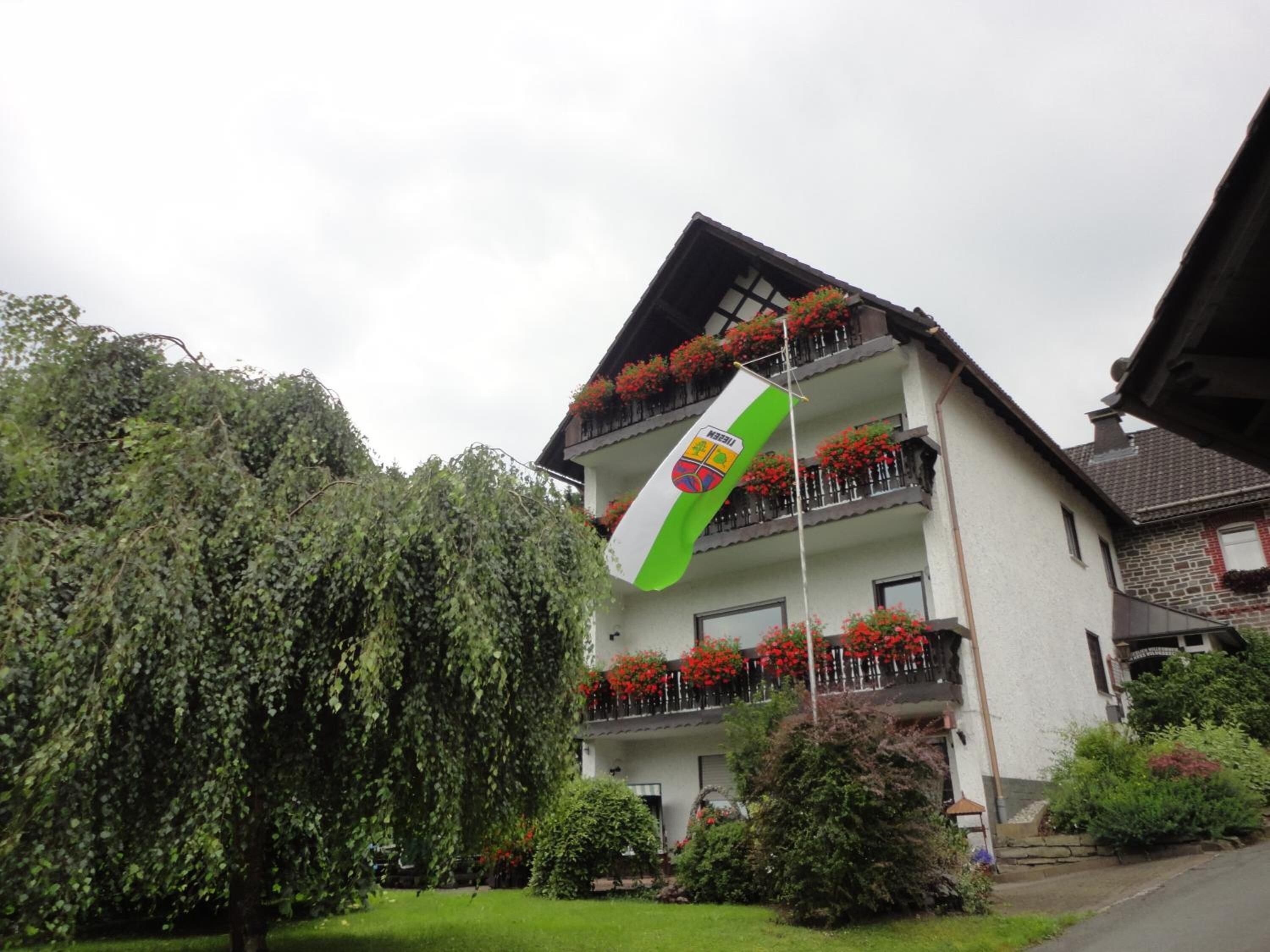 A multi-story building with balconies adorned with red flowers, a flag with a coat of arms, and a well-maintained garden.
