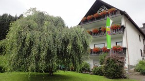 A building with a sloped roof, balconies with red flowers, and a large tree in the foreground. - Pension Haus Volkesbach (Hallenberg)