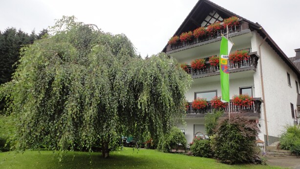 En byggnad med ett sluttande tak, balkonger prydda med röda blommor och ett stort träd i förgrunden.