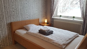 A neatly made bed with a geometric patterned bedspread, a pillow, and folded towels. There is a bedside table with a lamp and a small clock. The room has birch-patterned wallpaper and a window with curtains. - Villa Elsy (Vidsel)
