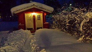 A small, snow-covered cabin with a red exterior and a white door, surrounded by snow.