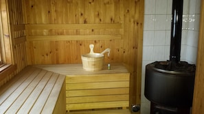 A sauna room with wooden walls, a wooden bench, a wooden bucket with a ladle, a small bottle, and a black heater. - Villa Elsy (Vidsel)