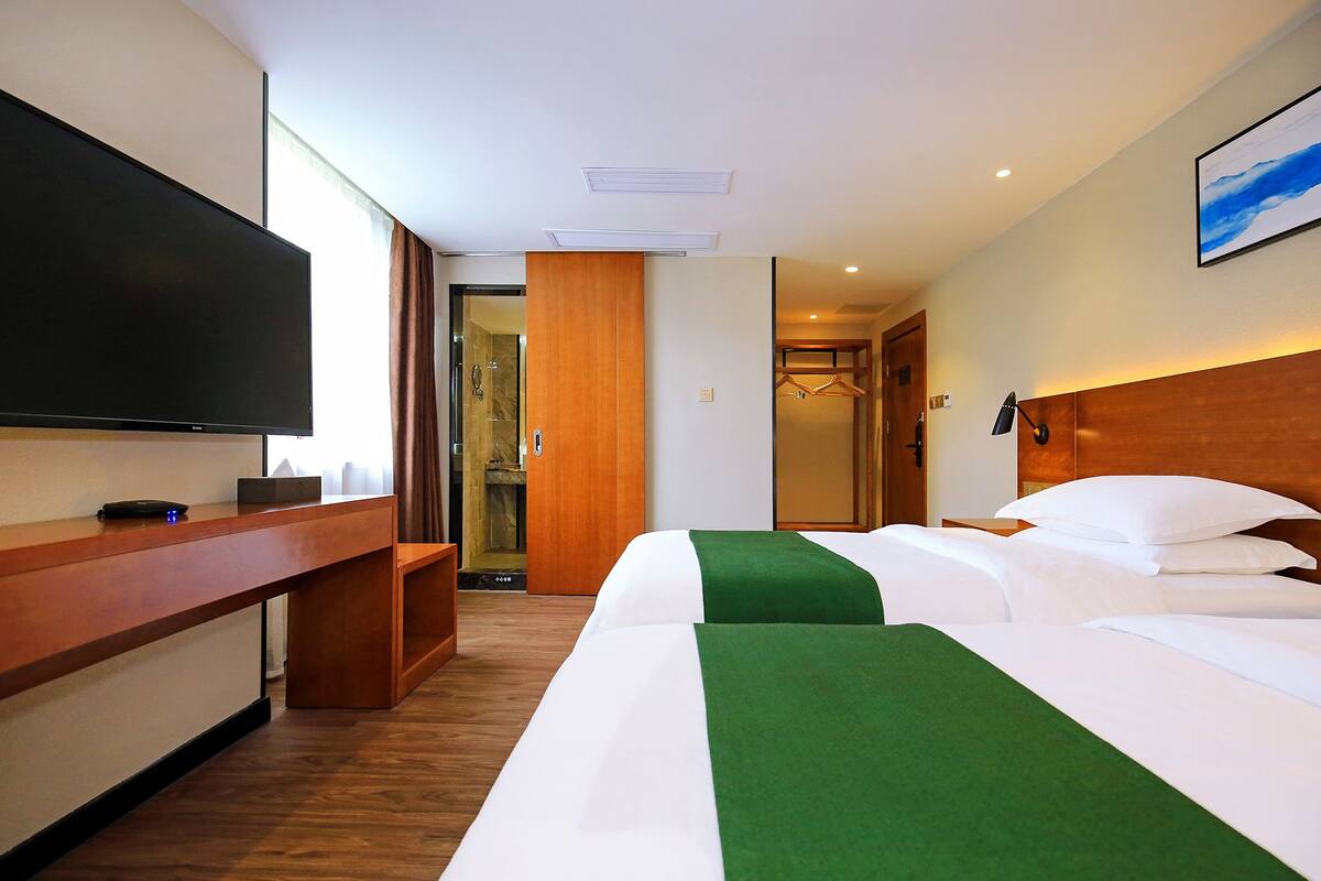 Habitación de hotel con un televisor de pantalla plana grande, un escritorio de madera, una cama con ropa de cama blanca y verde, y una zona de ducha.