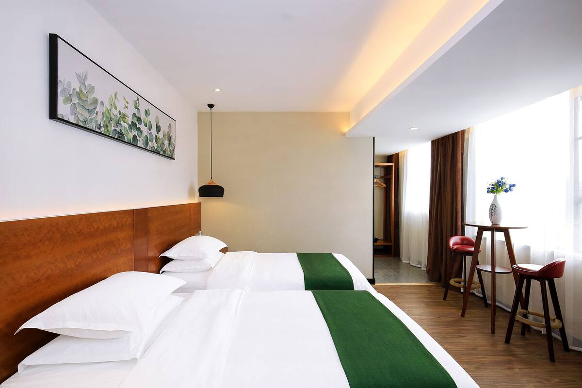 Habitación de hotel con dos camas, una mesita con sillas, un florero con flores y un cuadro en la pared.