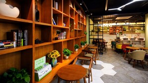 A cozy cafe with wooden shelves filled with books and decorative items, a seating area with round tables and chairs, and a visible QR code for digital payments.