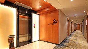 A hotel corridor with an elevator, a wall-mounted control panel, and a carpeted floor.