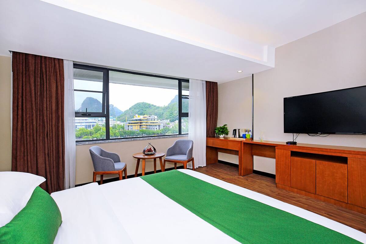 Habitación de hotel con una cama grande, dos sillas, una mesita, un escritorio, un televisor y una ventana con vistas a edificios y montañas.