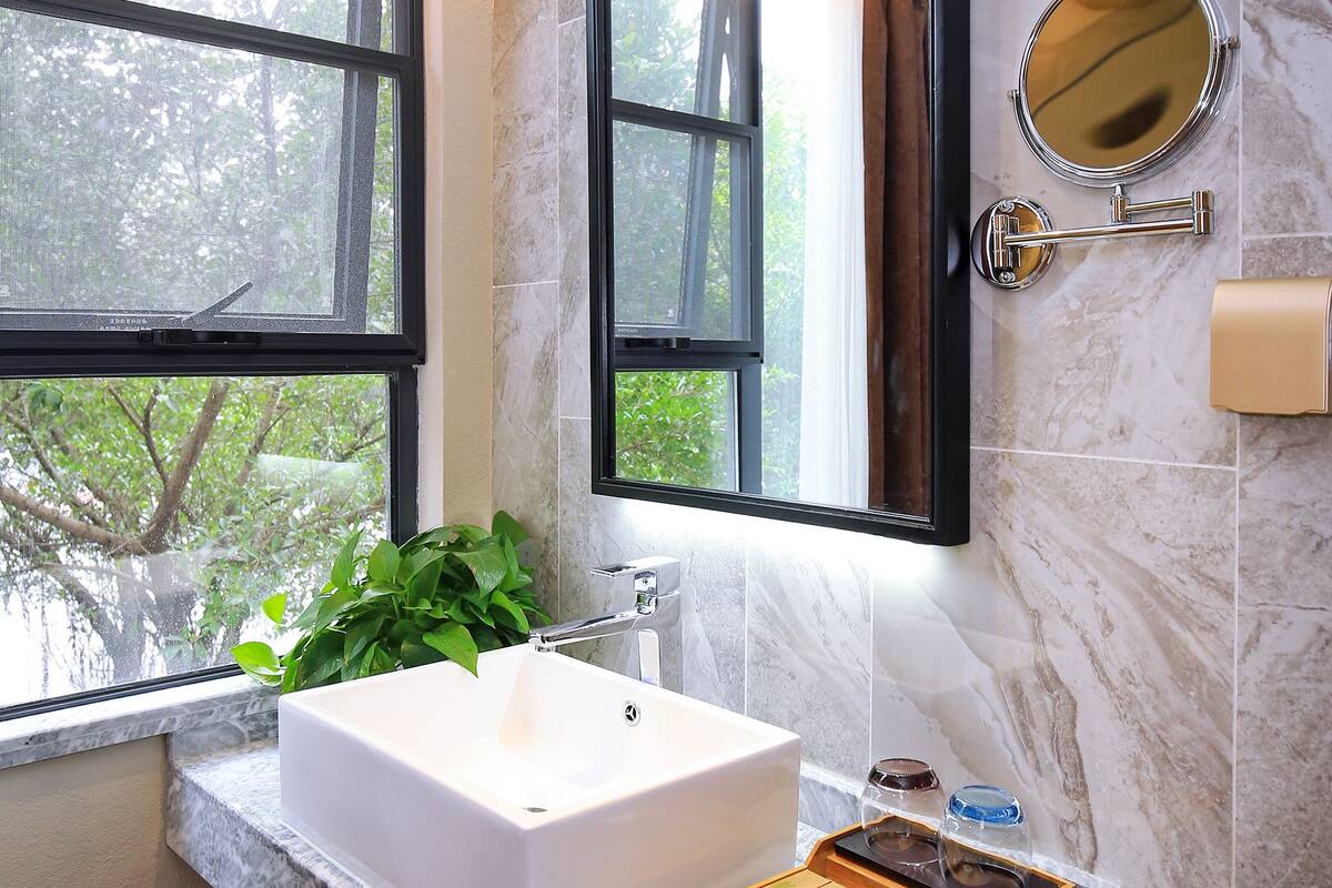 Un baño con un ventanal grande, un lavamanos blanco, un espejo y una bandeja de madera con artículos de aseo.
