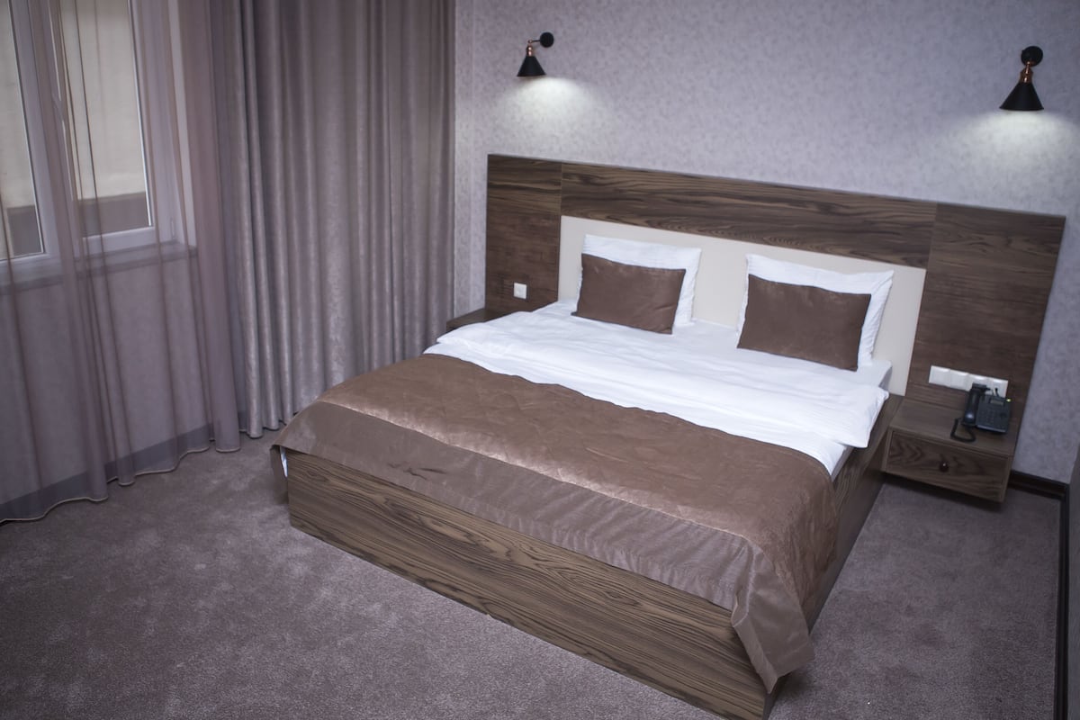 A neatly made bed with a wooden headboard, a bedside table with a phone, and a window with curtains.