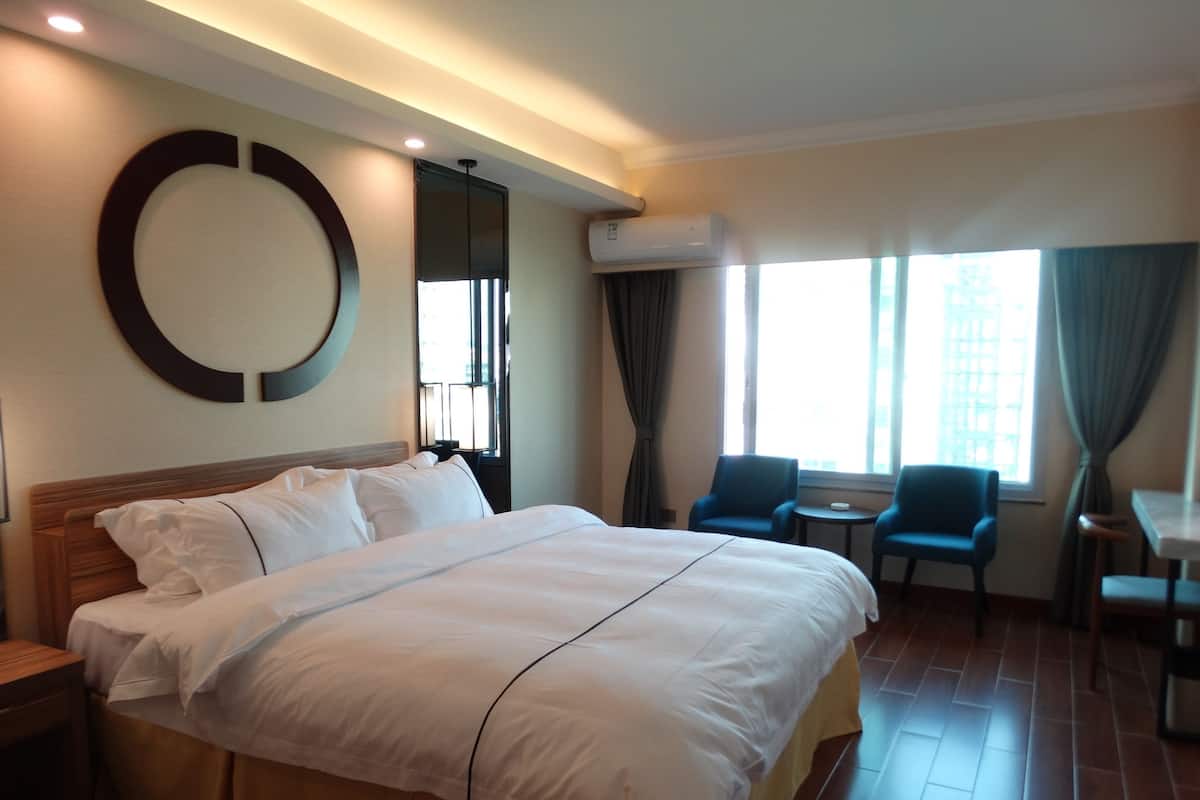 A hotel room with a large bed, a round mirror, a small table, two chairs, and a window with curtains.