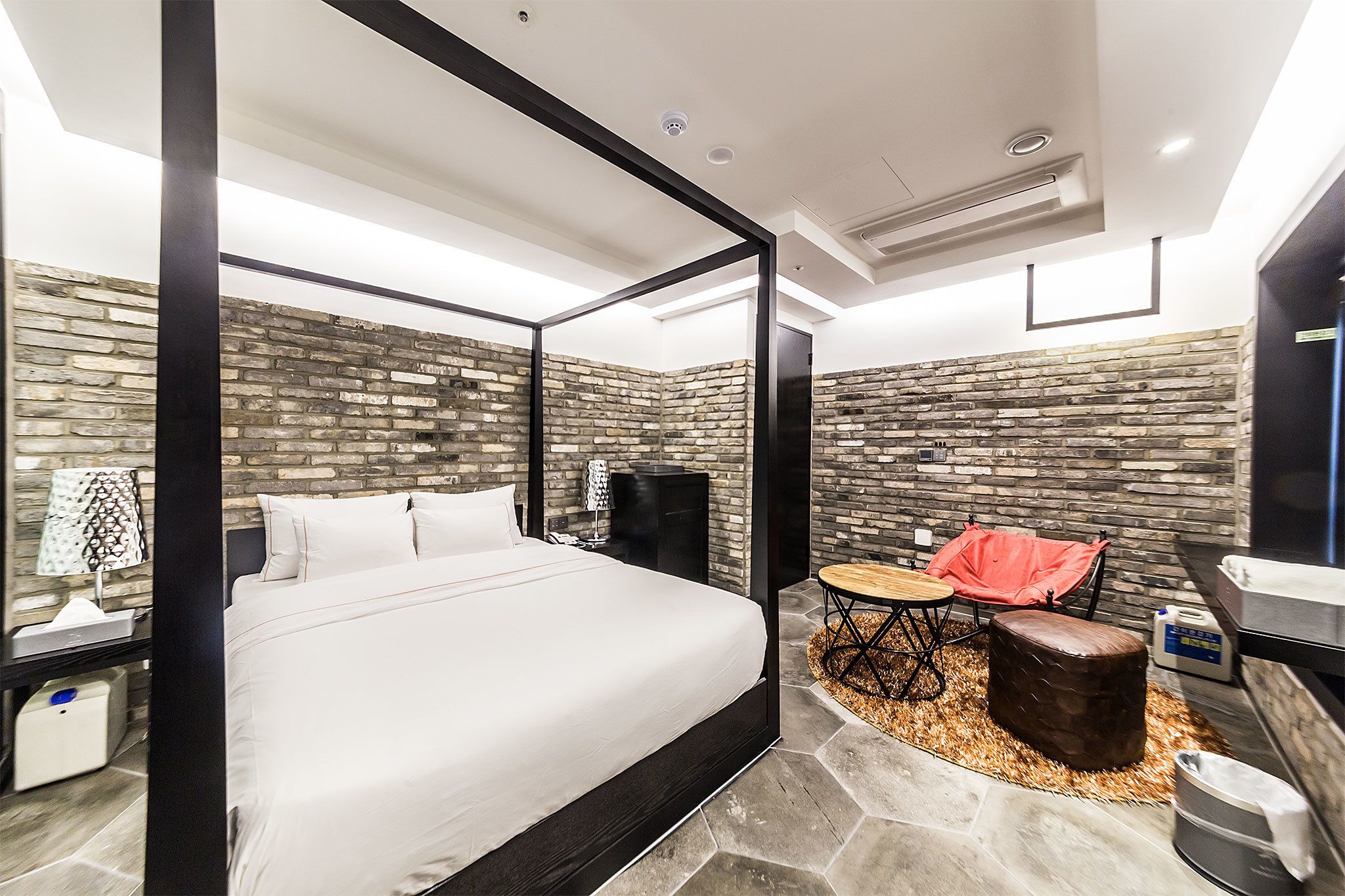 A modern hotel room with a four-poster bed, stone wall accents, a small round table with a chair, and a trash bin.