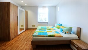 Une chambre avec un plancher en bois, un lit recouvert d’une couette aux couleurs vives, une armoire en bois, une fenêtre avec des stores et un radiateur.