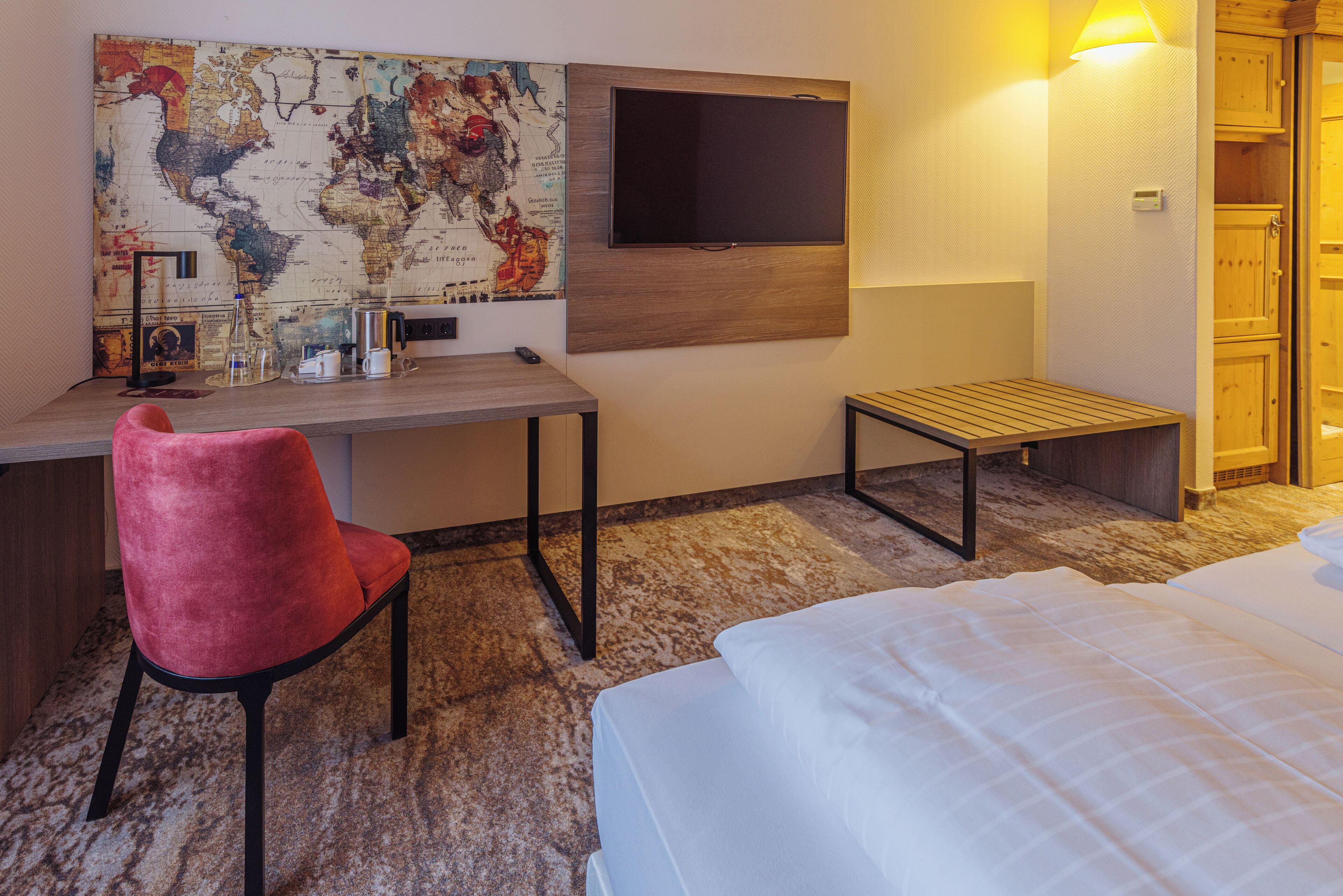 A hotel room with a bed, a desk with a chair, a TV mounted on the wall, and a wooden cabinet.