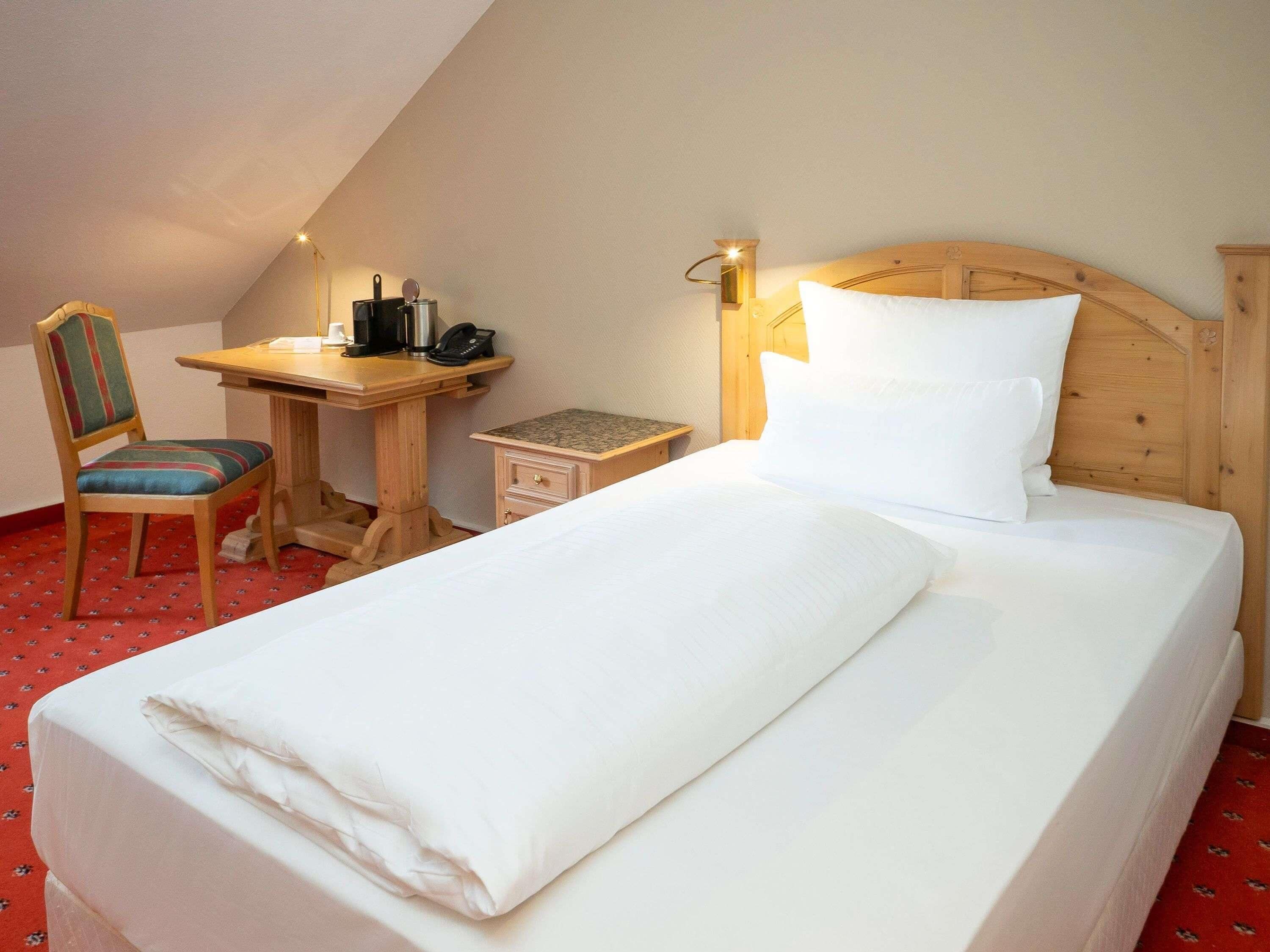 A single bed with a wooden headboard, a small desk with a chair, a nightstand, and a lamp.