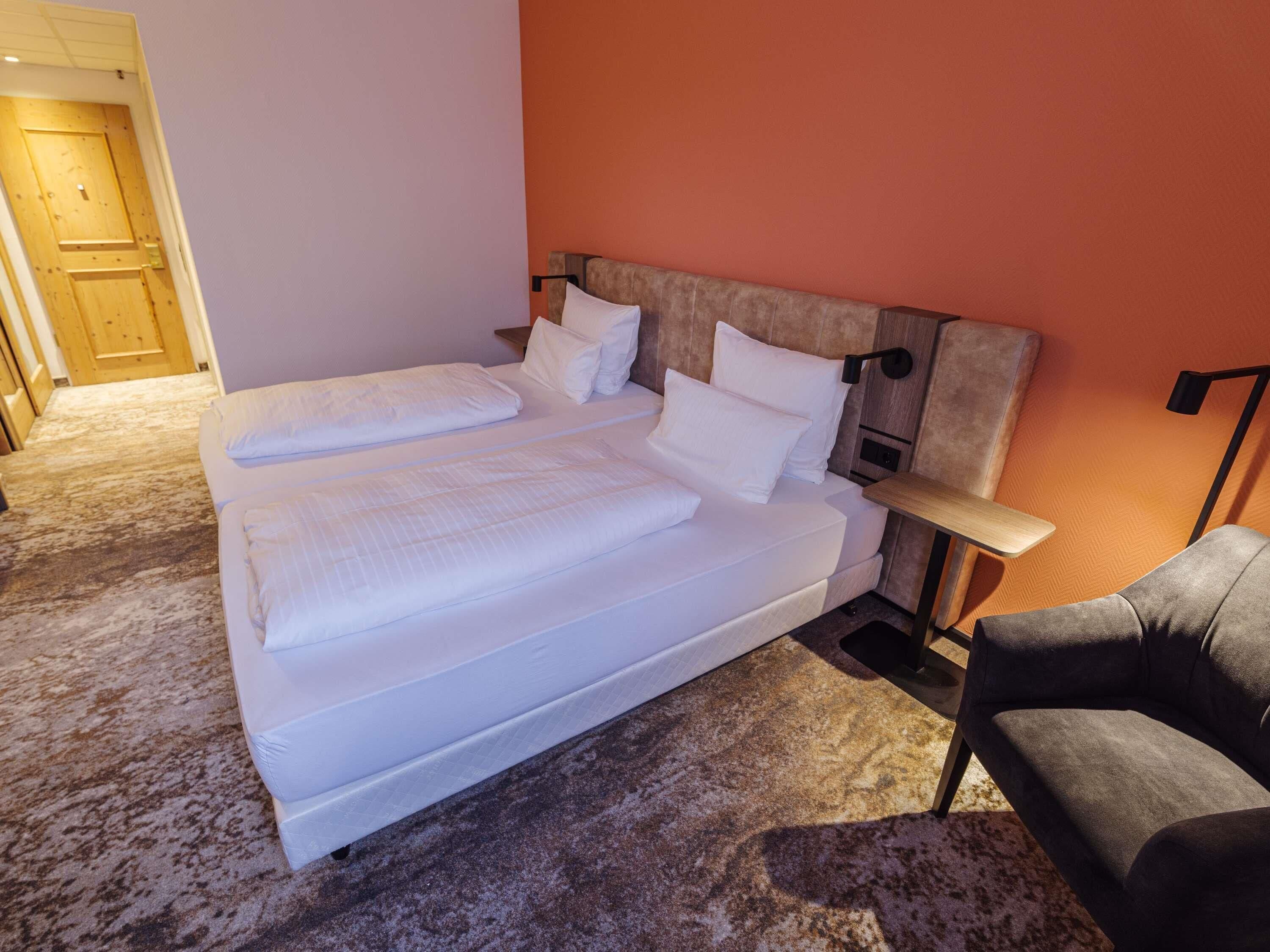A double bed with white linens, a bedside table, a chair, and a wooden door.