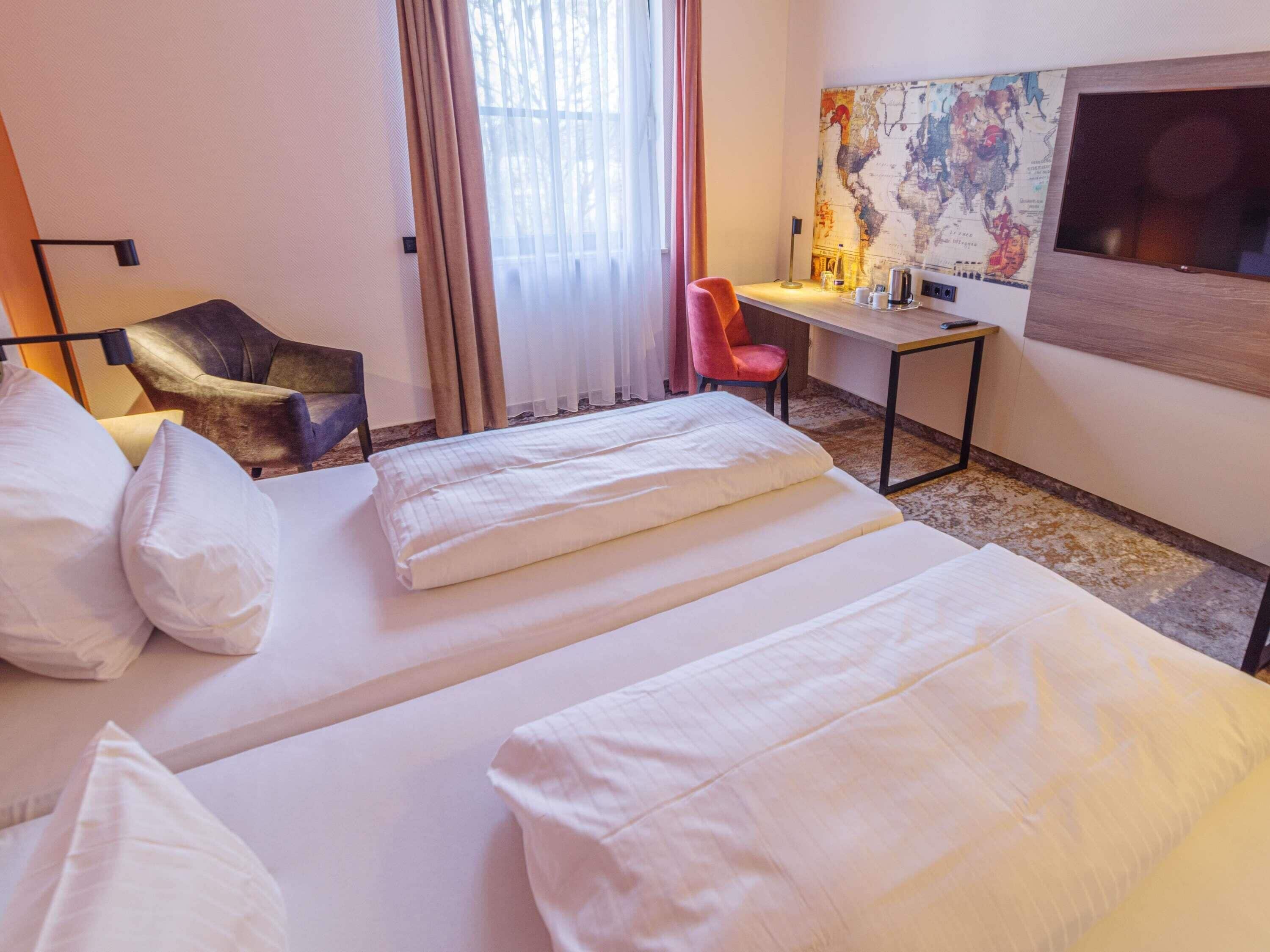 A hotel room with two beds, a desk, a chair, a map on the wall, and a window with curtains.