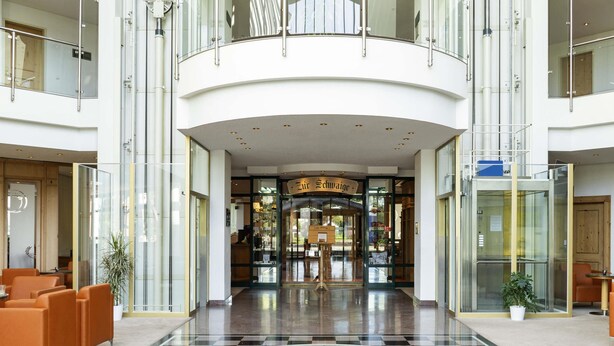 Eine geräumige Lobby mit Schachbrettmuster-Bodenbelag, Sitzbereichen und einem Glasgang zu einem Restaurant.
