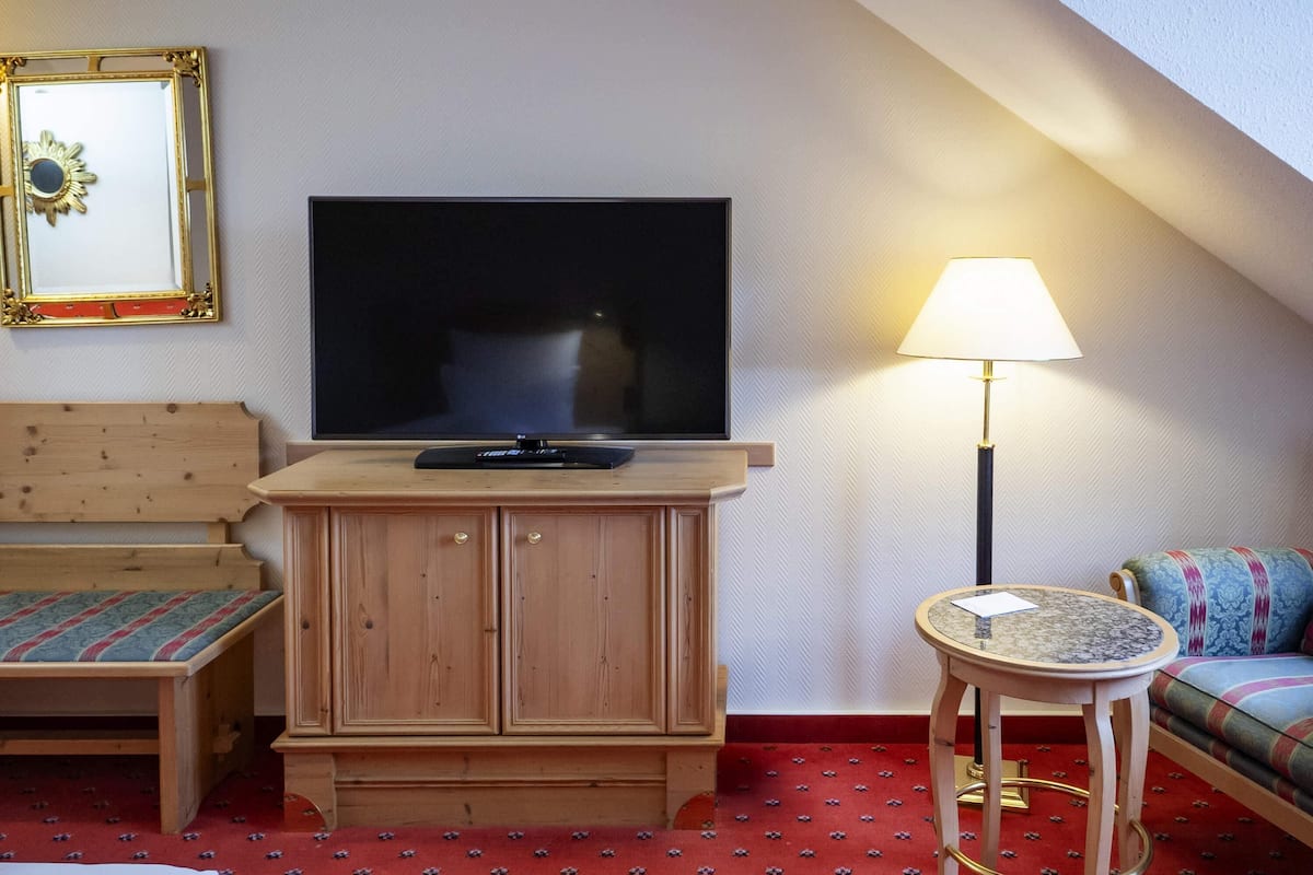Une piĂšce avec un tĂ©lĂ©viseur posĂ© sur un meuble en bois, un lit recouvert dâune courtepointe Ă motifs rouges, une petite table ronde avec une lampe et un canapĂ© agrĂ©mentĂ© de coussins Ă motifs.