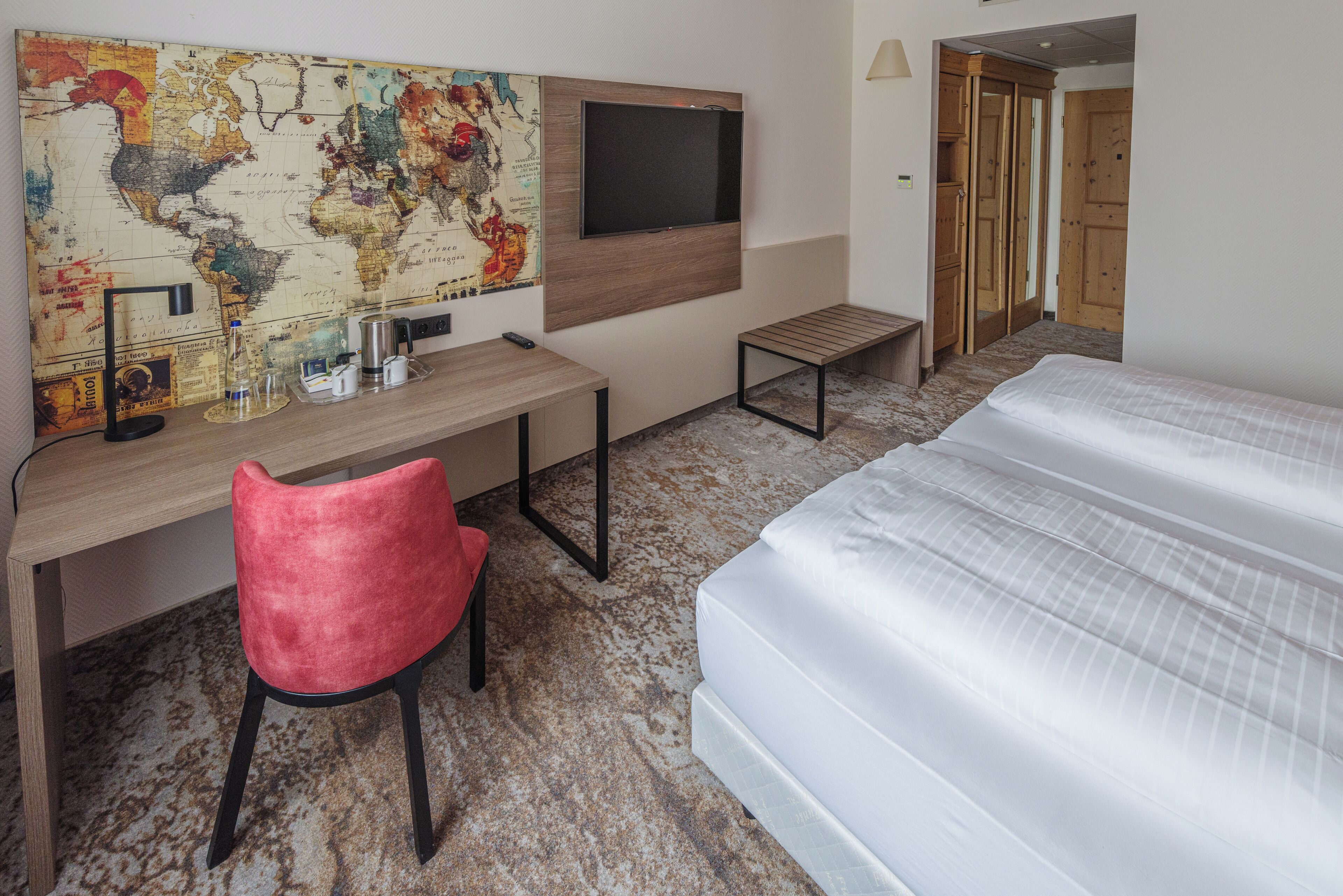 A hotel room with a bed, a desk with a chair, a map on the wall, and a TV.