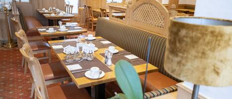 A restaurant with wooden tables and chairs, a patterned carpet, and a large potted plant.