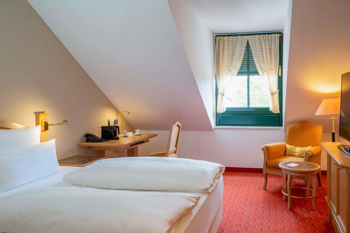 Ein Hotelzimmer mit Bett, Schreibtisch, Stuhl, einem kleinen Tisch und einem Fenster mit Vorhängen.