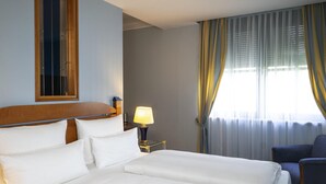 Ein Hotelzimmer mit einem großen Bett, einem Nachttisch mit Lampe, einem Sessel und einem Fenster mit Vorhängen.
