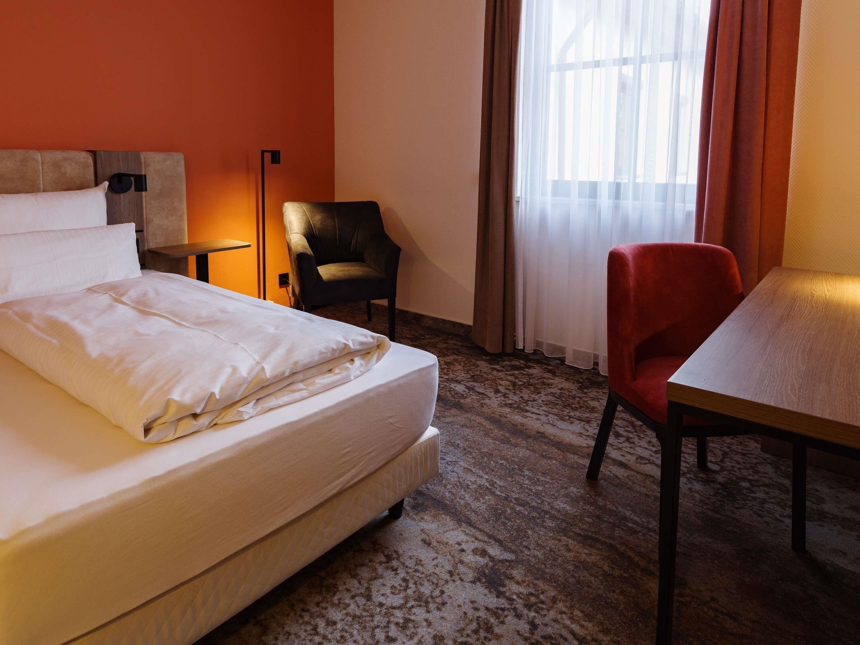 Une chambre d’hôtel avec un lit, un bureau, une chaise, une lampe et une fenêtre avec des rideaux.
