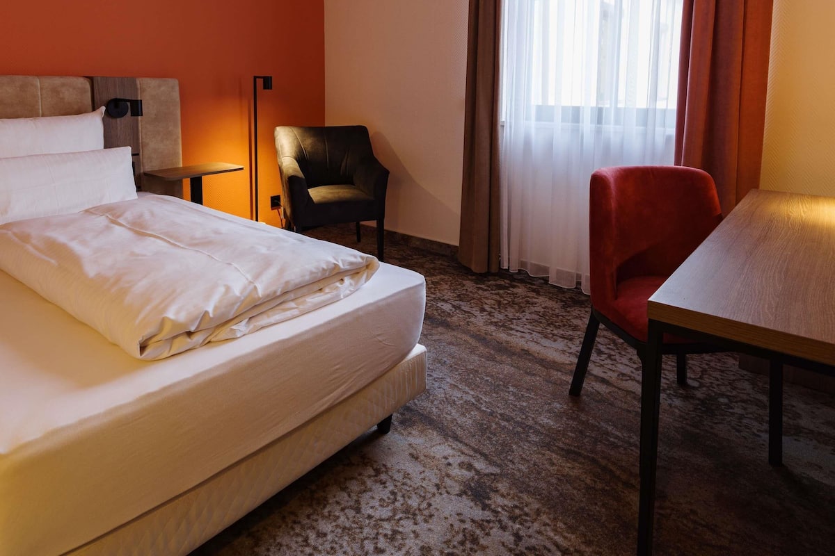 Une chambre d’hôtel avec un lit, un bureau, une chaise, une lampe et une fenêtre avec des rideaux.