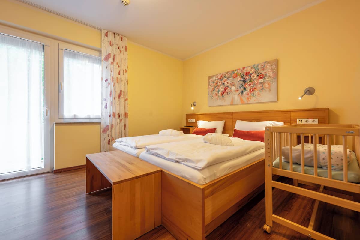 A bedroom with a wooden bed, a wooden bench, a crib, a painting on the wall, and a window with curtains.