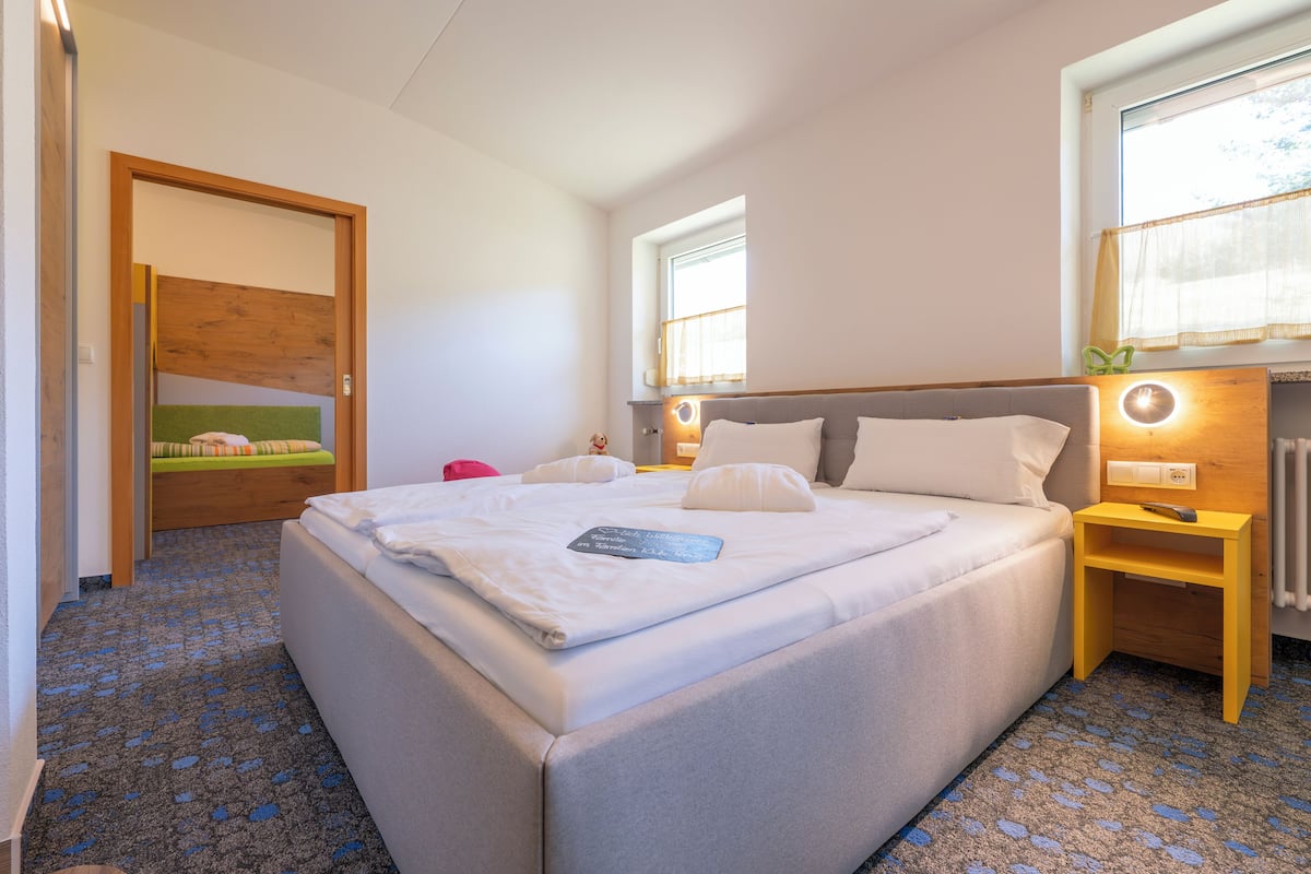 A hotel room with a large bed, a bedside table with a lamp, a window with curtains, and a wooden door.