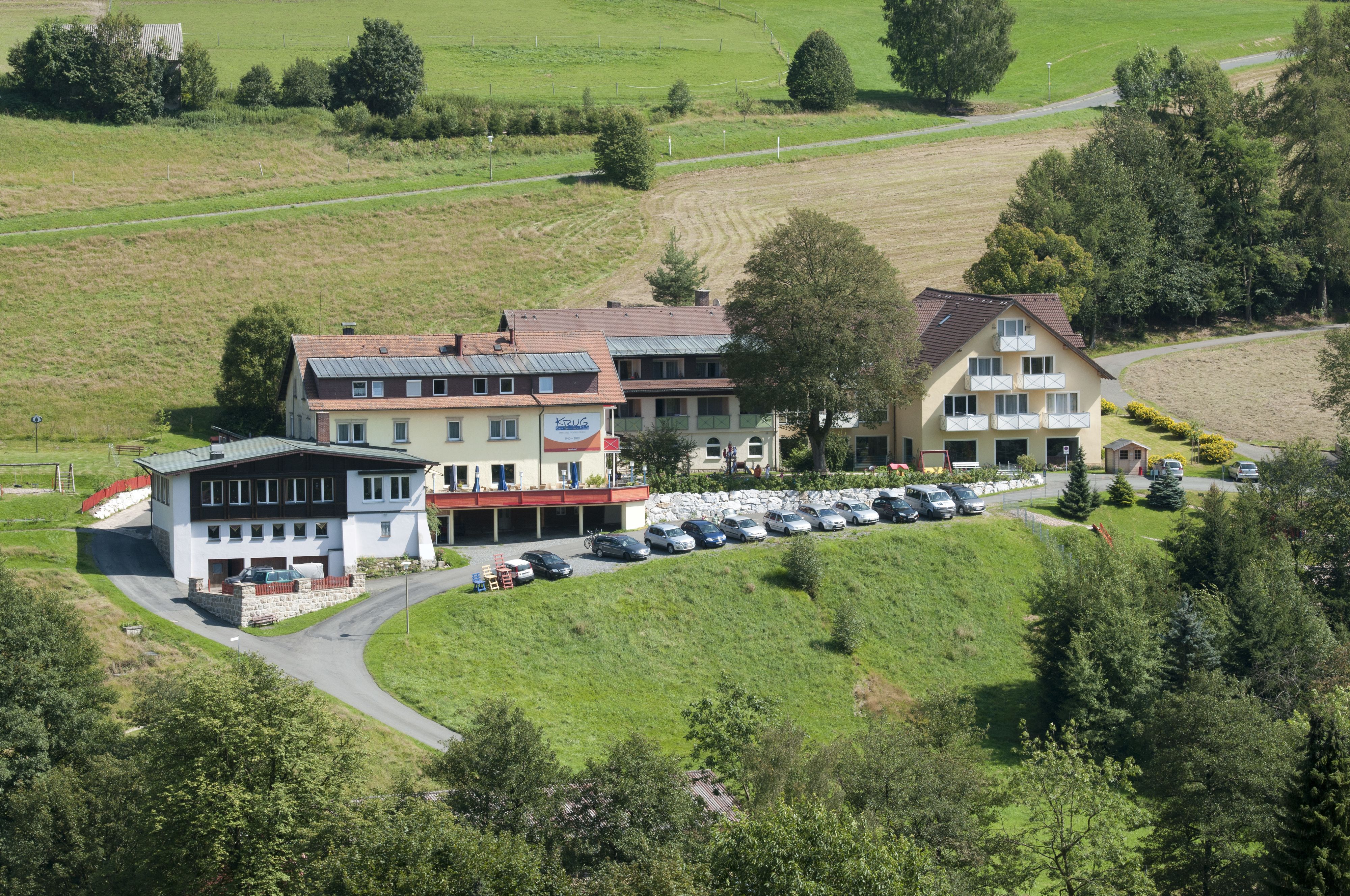 A large building with multiple floors, a parking lot with several cars, and a surrounding green landscape.