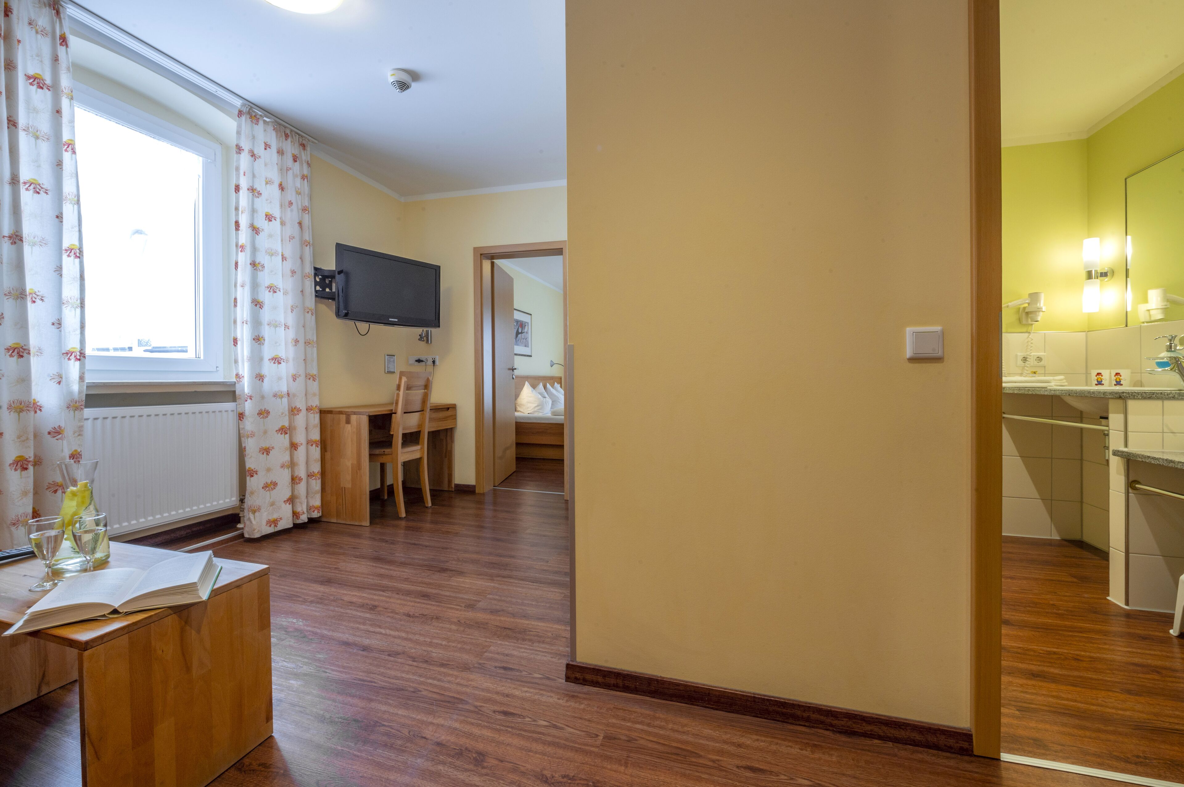A hotel room with a wooden floor, a desk with a chair, a TV mounted on the wall, and a bathroom with a sink and mirror.
