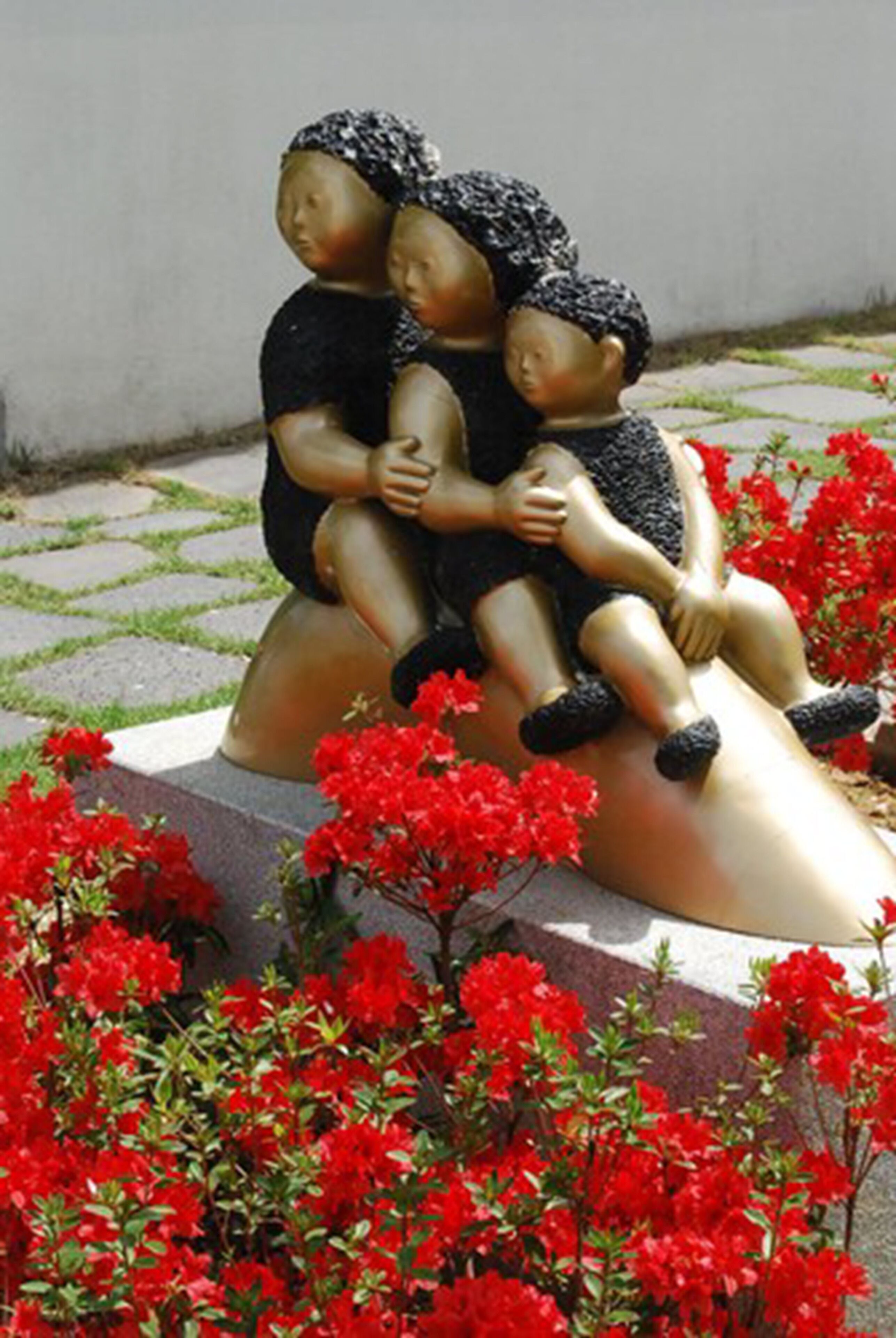 A sculpture of three figures seated on a curved structure surrounded by red flowers.