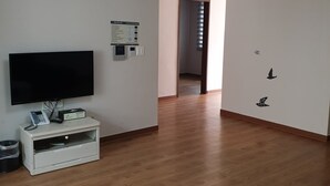 Ein geräumiges Zimmer mit einem Flachbildfernseher auf einem weißen Standfuß, einem kleinen Tischchen mit einem Telefon und einem Papierkorb. Der Boden ist mit Holz ausgelegt und es gibt eine Tür zu einem weiteren Bereich.