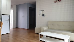 Ein modernes Wohnzimmer mit einem weißen Sofa, einem weißen Couchtisch und Holzboden. Im Hintergrund ist eine Küchenzeile mit Kühlschrank und Schränken zu sehen.
