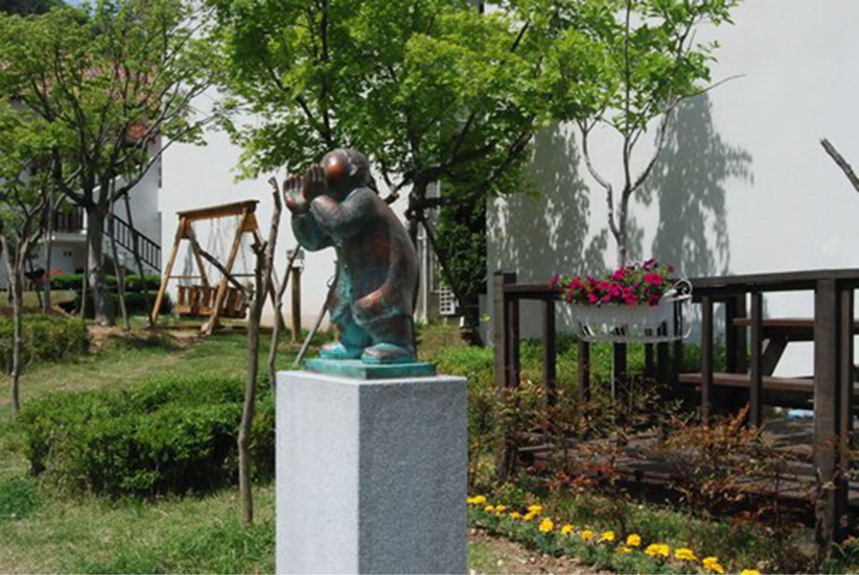 A statue of a person in a garden setting with a swing set and flowers in the background.