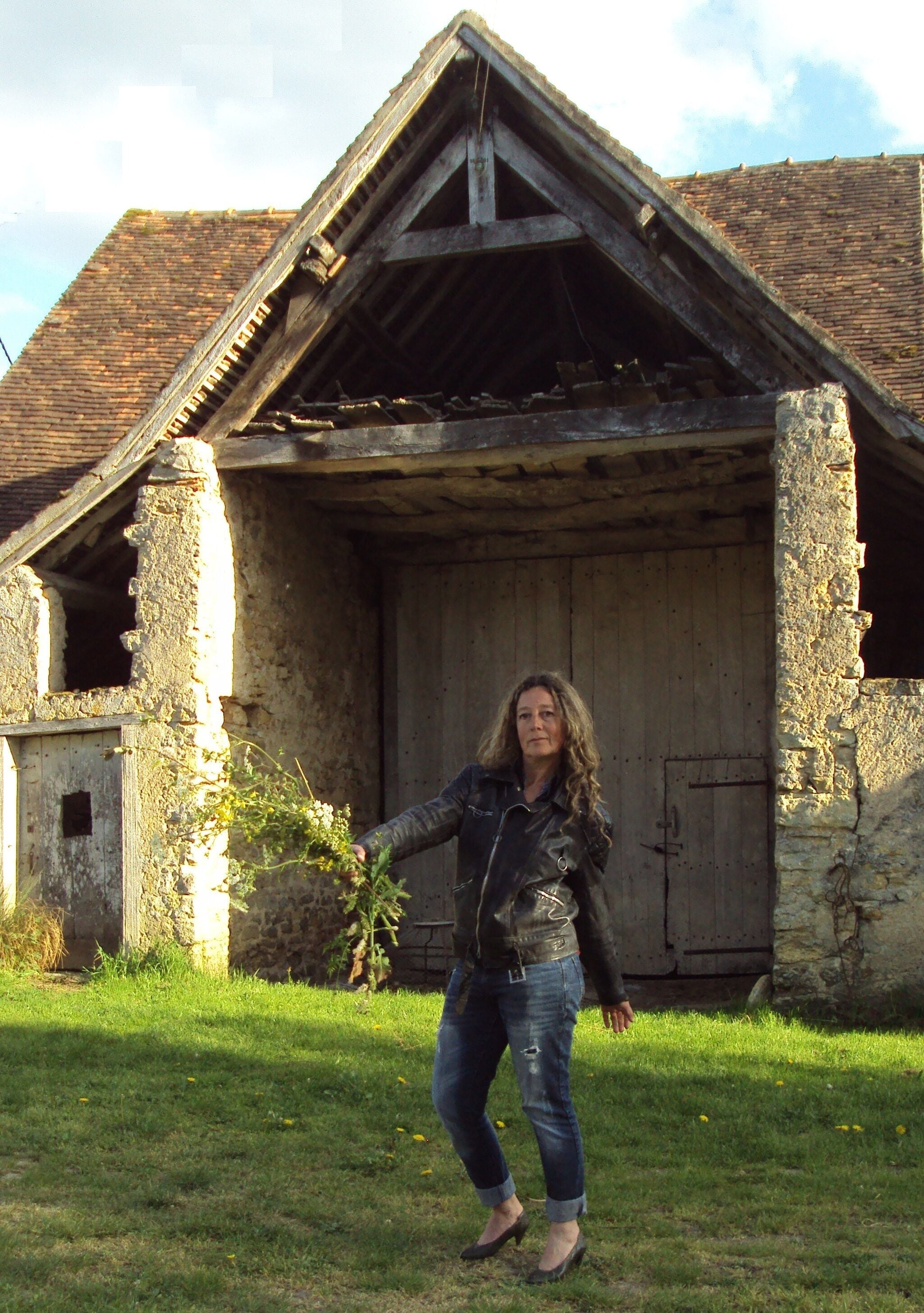 A woman standing in front of an old, rustic building with a sloped roof and stone walls.