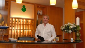 A hotel reception desk with a green logo, a vase of flowers, and a clock. - Hotel Sterkel (Roedermark)
