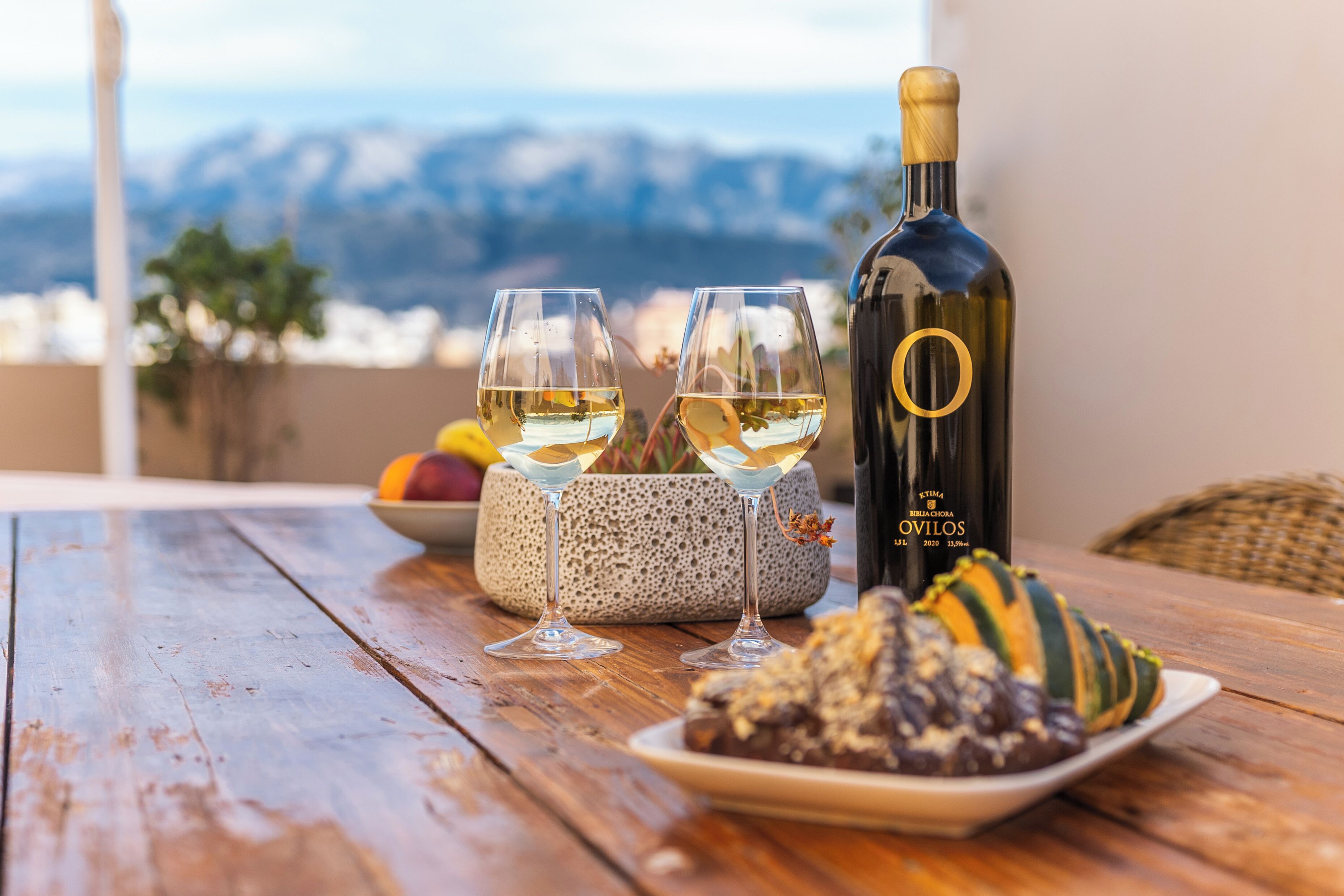 A wooden table with two wine glasses, a bottle of wine, and a plate of food on a balcony with a mountain view.