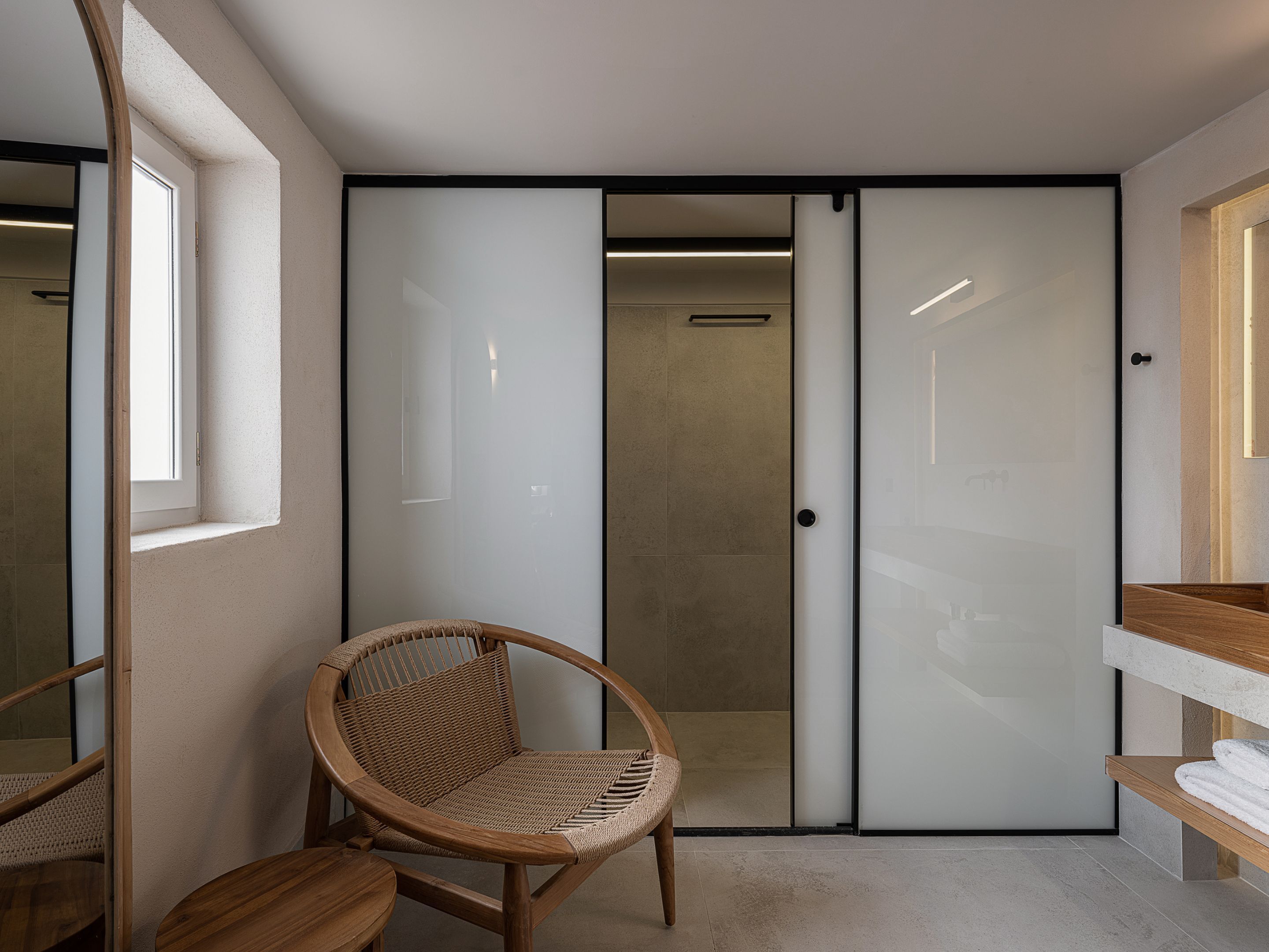 A modern bathroom with a glass shower enclosure, a wooden chair, and a wooden vanity with a towel rack.