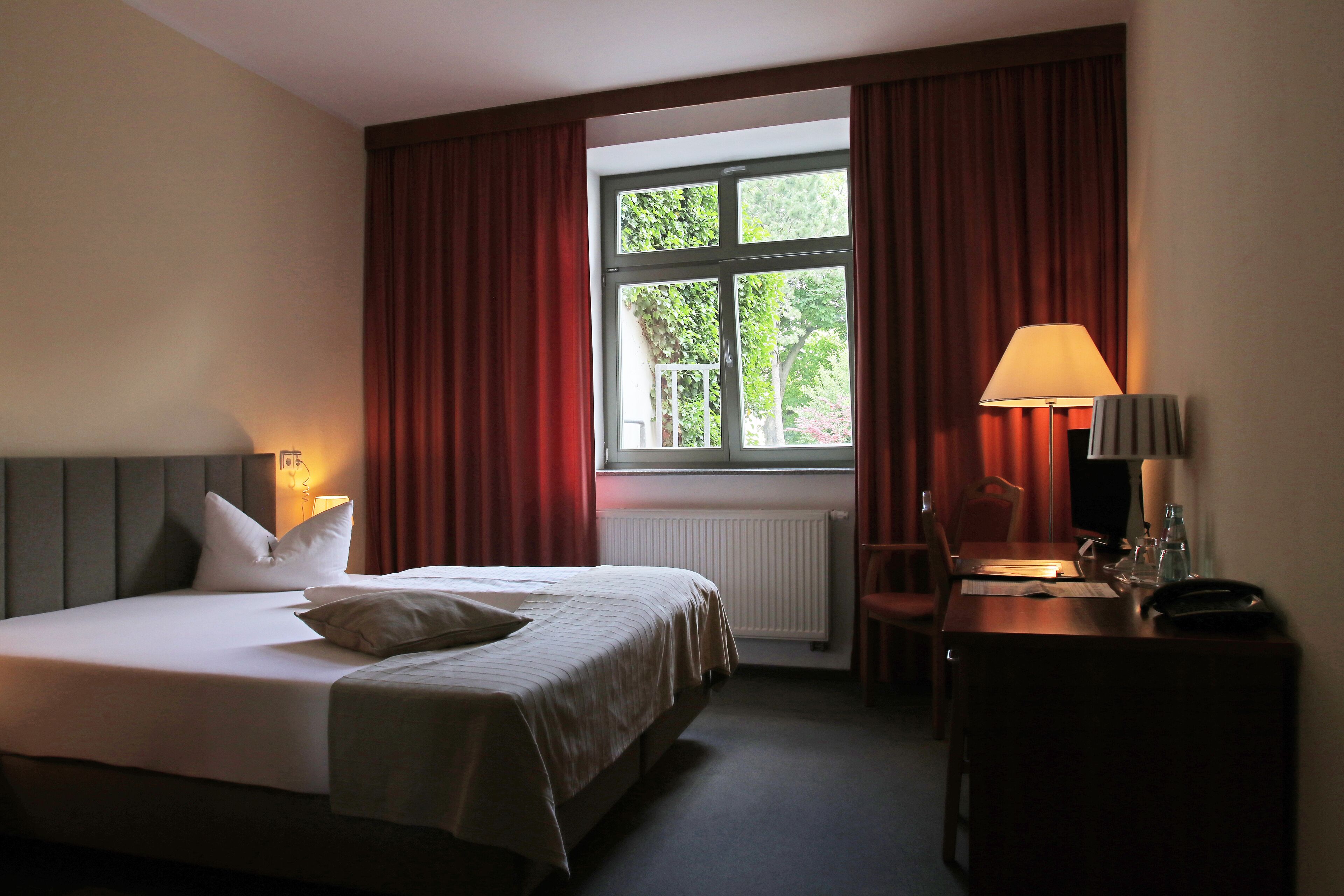 A hotel room with a bed, a desk, a chair, a lamp, and a window with curtains.