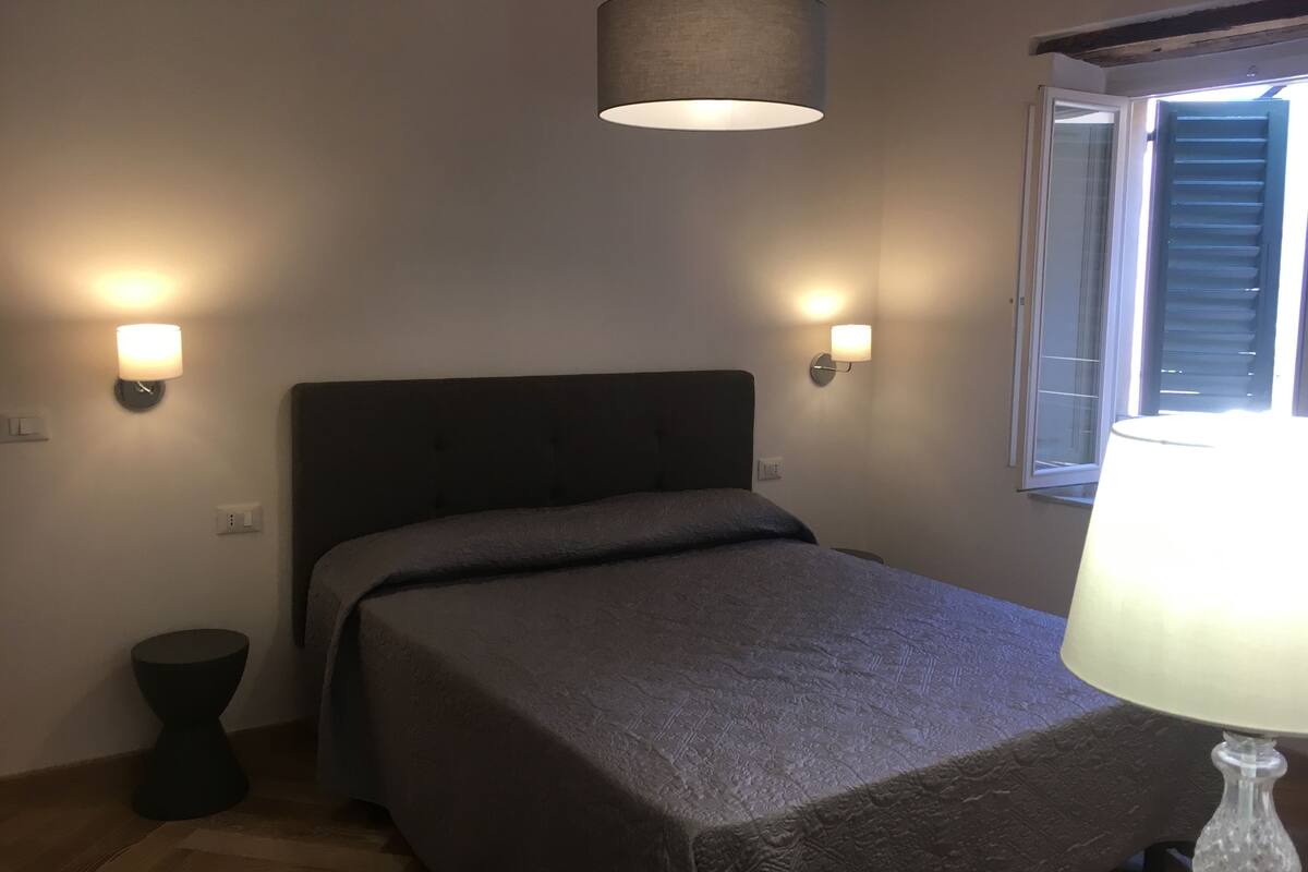 A bedroom with a bed, two wall-mounted lamps, a window with blinds, and a hanging lamp.