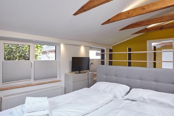 A modern bedroom with a bed, a TV, and a window with blinds. - Ferienhäuser & Appartments Düneneck 6 (St. Peter-Ording)