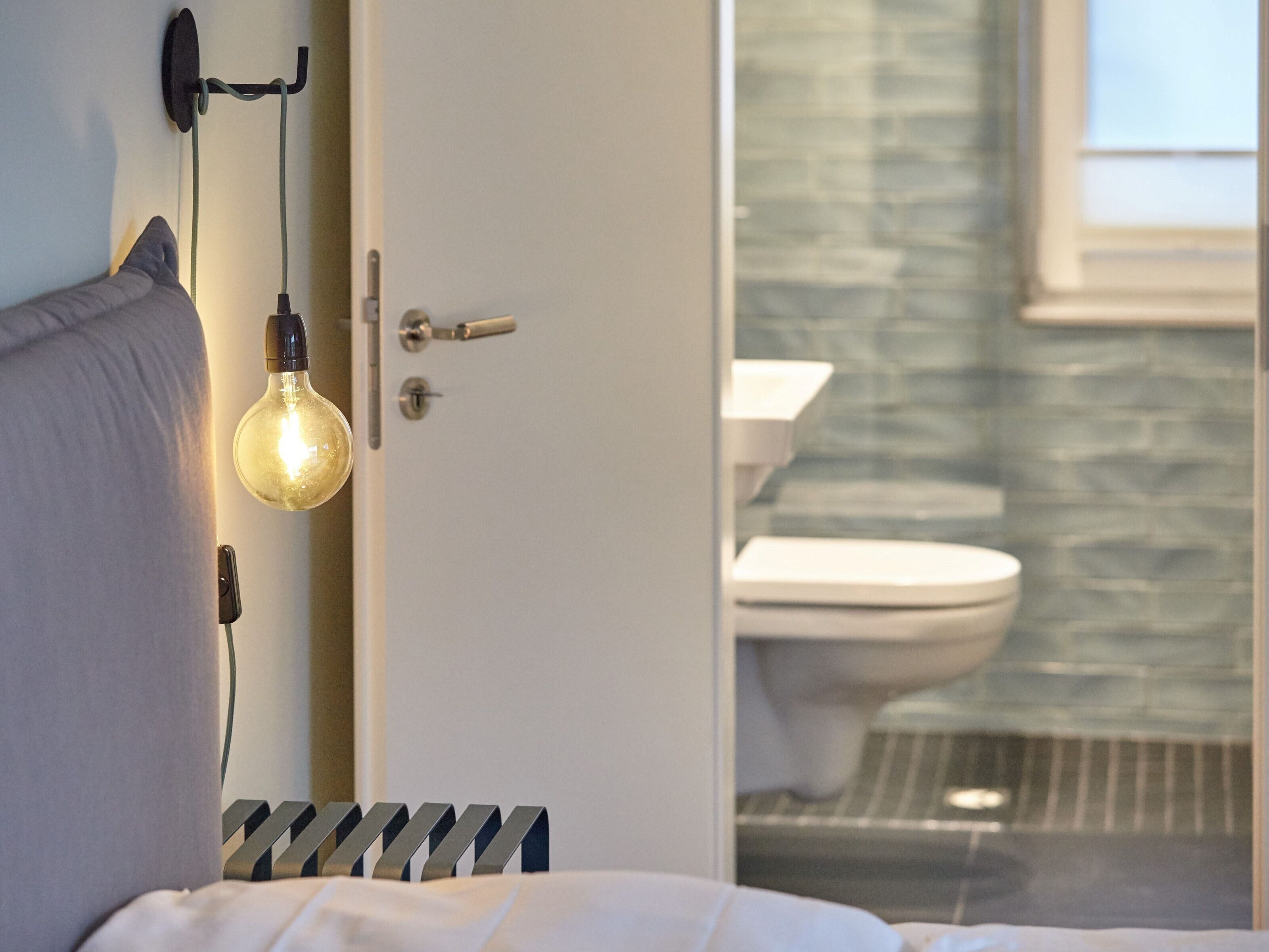 Quarto com cama, luminária de cabeceira e porta para banheiro com vaso sanitário e chuveiro.